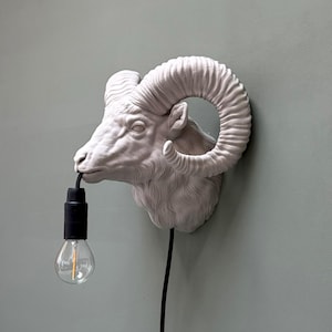 May include: A white ram head wall sconce with a black cord and a lightbulb. The ram head is mounted on a light green wall. The lightbulb is clear with a visible filament.