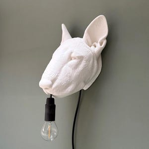May include: A white, textured Bull Terrier head wall lamp with a black cord and bulb socket. The lamp features a clear light bulb. The dog head is mounted on a green wall.