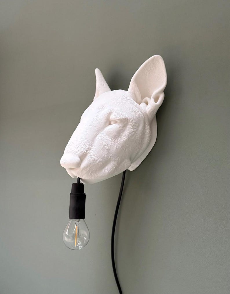 May include: A white, textured Bull Terrier head wall lamp with a black cord and bulb socket. The lamp features a clear light bulb. The dog head is mounted on a green wall.
