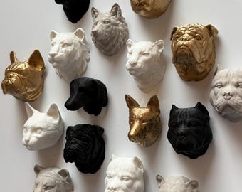Puede incluir: Una colección de adornos de pared con forma de cabeza de animal en varios colores, incluyendo dorado, negro, blanco y gris. La colección presenta una variedad de animales, como gatos, perros y leones. Las cabezas están esculpidas con detalles y montadas sobre una superficie blanca.