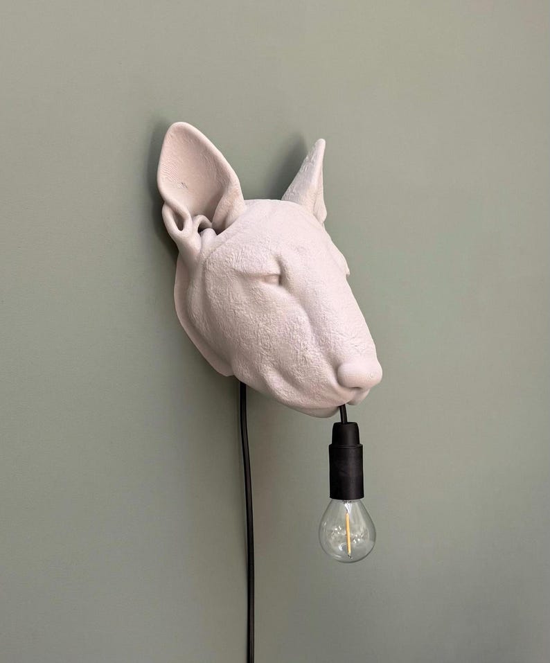 May include: A light-colored, dog-shaped wall lamp with a black cord and a lightbulb hanging from its mouth. The lamp is mounted on a light green wall. The dog's head is the focal point of the design.
