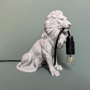 Könnte beinhalten: Eine weiße Keramik-Löwenfigur mit einem schwarzen Kabel und einer Glühbirne im Maul. Der Löwe sitzt auf einer hellgrünen Oberfläche.