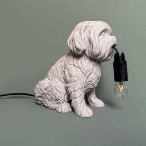 Può includere: Una scultura di cane in ceramica bianca con una lampadina in bocca. Il cane è seduto su una superficie verde e ha un cavo nero attaccato alla lampadina.