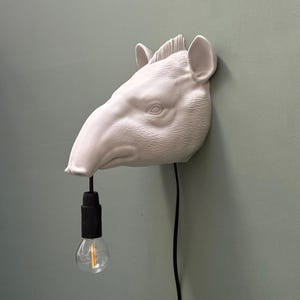 Peut inclure: Une applique murale blanc mat en forme de tête de tapir. La sculpture animale présente un long museau, des oreilles pointues et une courte crinière. Un cordon électrique noir pend du museau, se terminant par une douille noire et une ampoule.