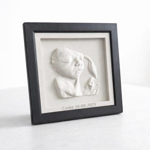 Könnte beinhalten: Gerahmte weiße Reliefskulptur eines Babygesichts und einer Hand. Die Skulptur befindet sich vor einem weißen Hintergrund in einem schwarzen Rahmen. Der Text "Lorèn 16-08-2025" ist unten eingeschrieben.