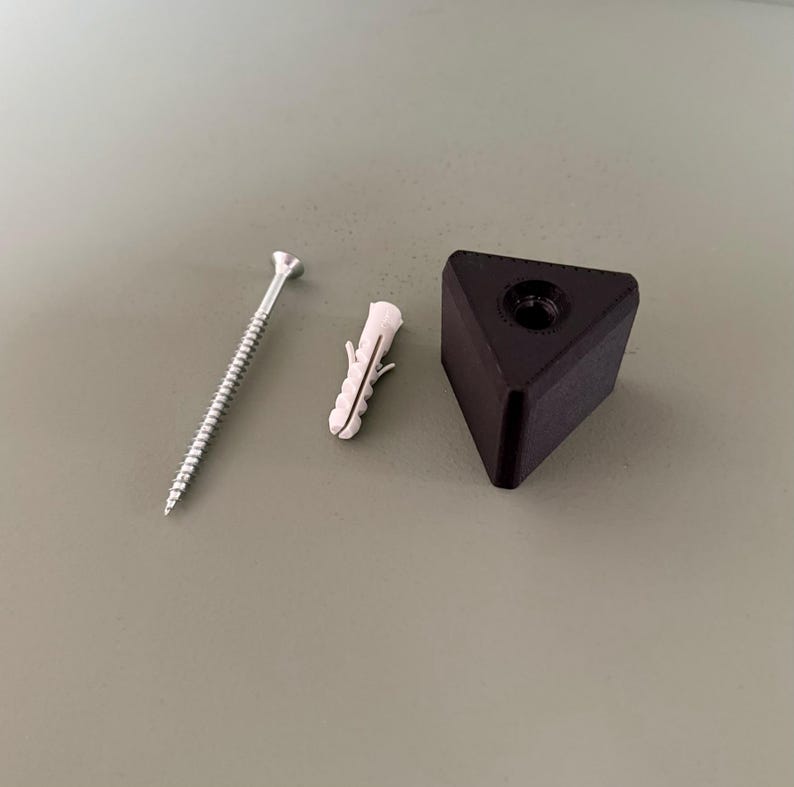 May include: A set of furniture hardware components, including a silver screw, a white plastic wall anchor, and a black triangular furniture foot with a threaded insert. The items are arranged on a neutral-toned surface.