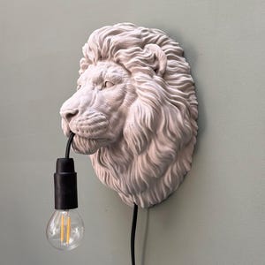 Op de afbeelding: Een witte leeuwenkop wandlamp met een zwarte lampfitting en een heldere gloeilamp. De manen van de leeuw zijn gedetailleerd en gestructureerd. De lampfitting hangt uit de bek van de leeuw. De achtergrond is een gedempt groen.