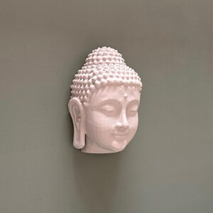 May include: A white Buddha head sculpture with detailed facial features and a textured, rounded hairstyle. The sculpture is mounted on a muted green wall, creating a serene and minimalist aesthetic. The head is angled slightly, showcasing the serene expression.
