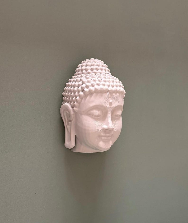 May include: A white Buddha head sculpture with detailed facial features and a textured, rounded hairstyle. The sculpture is mounted on a muted green wall, creating a serene and minimalist aesthetic. The head is angled slightly, showcasing the serene expression.