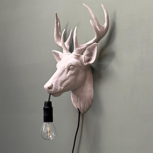 Puede incluir: Una escultura de pared con forma de cabeza de ciervo en color blanco mate, con un cable eléctrico negro que sale de su boca. El cable se conecta a un casquillo negro que sostiene una bombilla de cristal transparente con filamento visible.