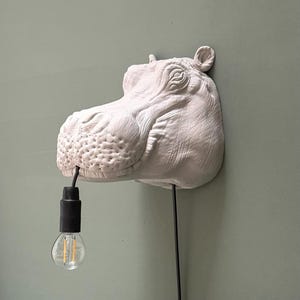 Può includere: Una lampada da parete a forma di testa di ippopotamo scolpita in bianco. L'ippopotamo presenta una texture della pelle dettagliata e piccole orecchie. Un cavo elettrico nero esce dalla bocca, sostenendo una lampadina in vetro trasparente.