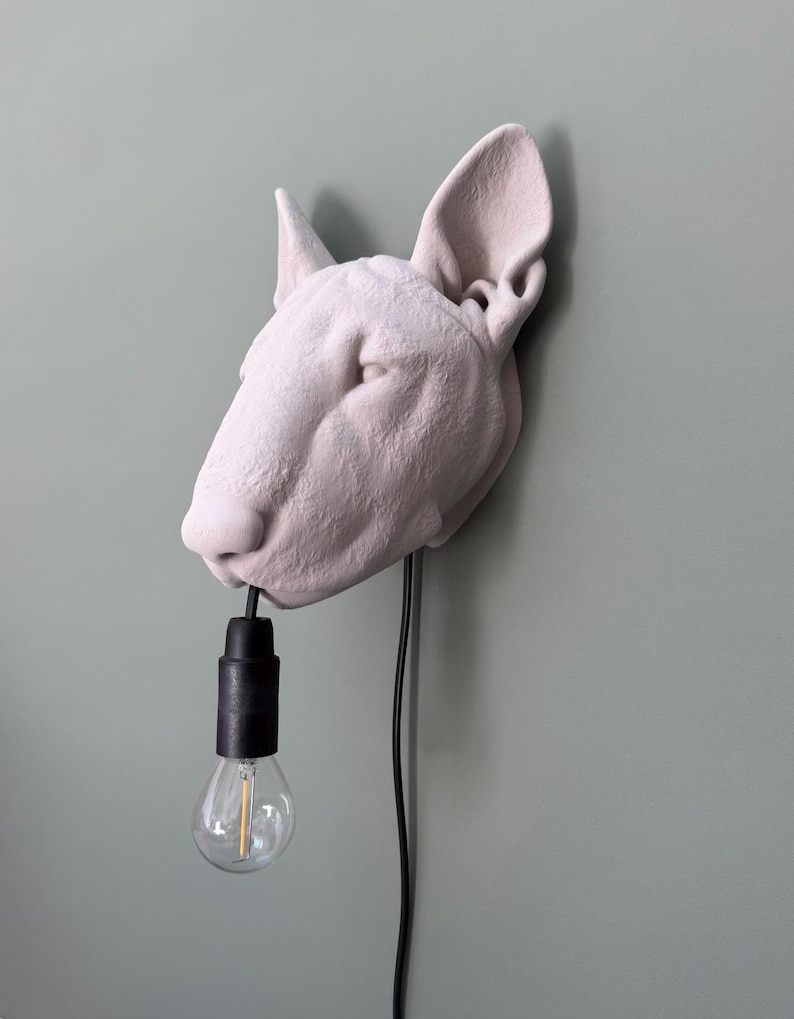 May include: A light-colored, dog-shaped wall lamp with a black bulb socket and a clear light bulb. The lamp is mounted on a light green wall, with a black power cord extending down. The dog's head is the focal point.