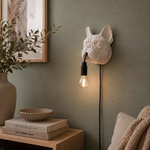 Può includere: Una lampada da parete bianca a forma di testa di bulldog francese. La bocca del cane sostiene un cavo nero con una singola lampadina in vetro trasparente che pende verso il basso. La lampada è montata a parete e ha un cavo di alimentazione nero.