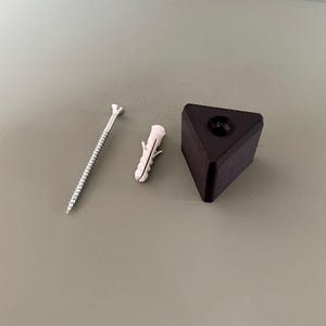 May include: A close-up of hardware components, including a silver screw, a white plastic wall anchor, and a black triangular furniture foot with a threaded insert. The items are arranged on a neutral-toned surface.
