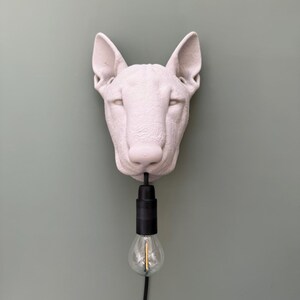 May include: A white, sculptural dog head wall lamp with a black light fixture and a clear lightbulb. The dog's head is mounted on a light green wall.