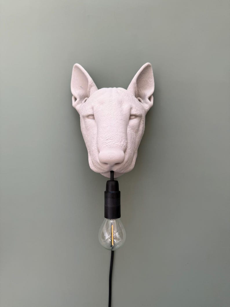 May include: A white, sculptural dog head wall lamp with a black light fixture and a clear lightbulb. The dog's head is mounted on a light green wall.