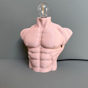 Puede incluir: Una lámpara de torso humano esculpido de color rosa con una bombilla de vidrio transparente. La lámpara está sobre una superficie gris.