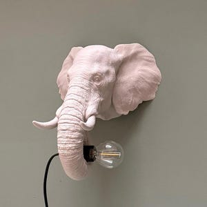 May include: A white elephant head wall sconce with a light bulb. The elephant head is detailed with wrinkles and texture. The light bulb is clear with a visible filament. A black cord extends from the elephant's trunk.