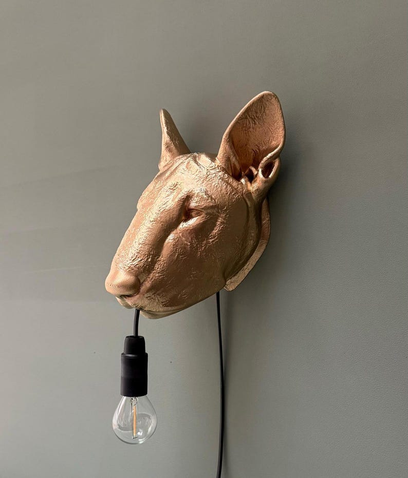 May include: A gold-colored Bull Terrier head wall lamp with a black cord and a clear lightbulb. The dog head is mounted on a wall, creating a unique lighting fixture. The lamp is a decorative piece.