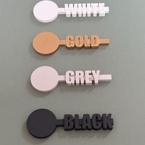 May include: Four color-coded, 3D-printed color swatches. Each swatch features a circular handle and a rectangular section with a color name. Colors include white, gold, grey, and black.