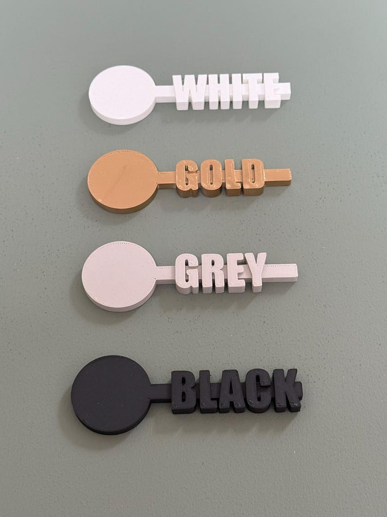 May include: Four color-coded, 3D-printed color swatches. Each swatch features a circular handle and a rectangular section with a color name. Colors include white, gold, grey, and black.
