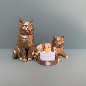 May include: Two gold cat ornaments, one sitting and one lying down, either side of a small candle holder with a battery-powered candle. The cat sculptures have detailed features and a metallic finish, creating a decorative accent.