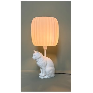 Könnte beinhalten: Eine weiße Tischlampe in Katzenform mit einem strukturierten, zylindrischen Lampenschirm. Die Lampe hat einen Sockel in Form einer sitzenden Katzenfigur und einen vertikal gerippten Lampenschirm. Die Lampe ist mit einem schwarzen Kabel an eine Steckdose angeschlossen. Die Lampe steht auf einer grauen Oberfläche.