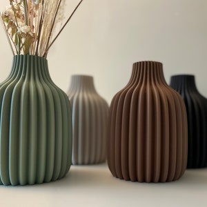 May include: Four decorative vases in green, beige, brown, and black. The vases have a ribbed design and are arranged on a white surface. The green vase holds dried flowers.