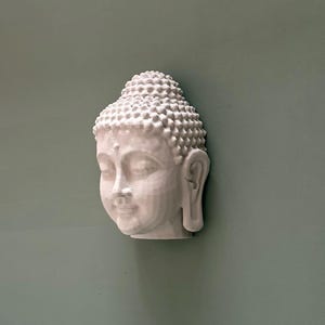 May include: A white, three-dimensional Buddha head sculpture with detailed facial features and a textured, rounded hairstyle. The sculpture is mounted on a muted green wall, creating a serene and minimalist aesthetic. The Buddha's serene expression and intricate details are highlighted.