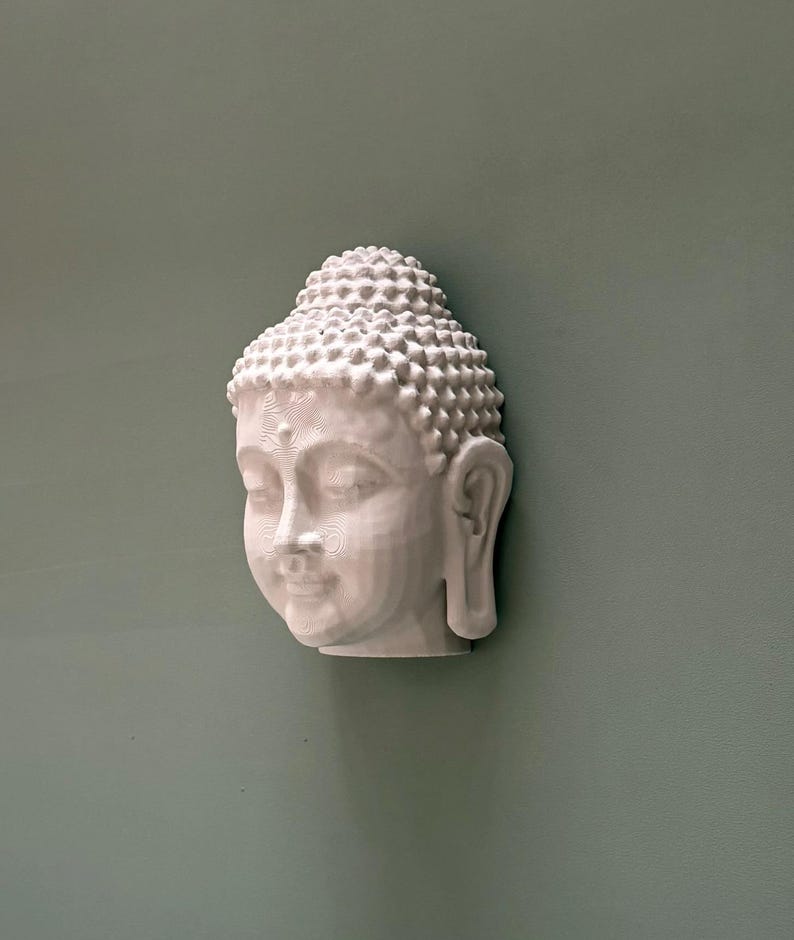 May include: A white, three-dimensional Buddha head sculpture with detailed facial features and a textured, rounded hairstyle. The sculpture is mounted on a muted green wall, creating a serene and minimalist aesthetic. The Buddha's serene expression and intricate details are highlighted.