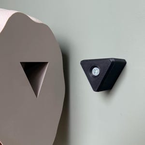 May include: A close-up of a gray, abstract-shaped object with a triangular cutout, next to a black, triangular object with a screw in the center. Both objects are mounted on a light green wall.
