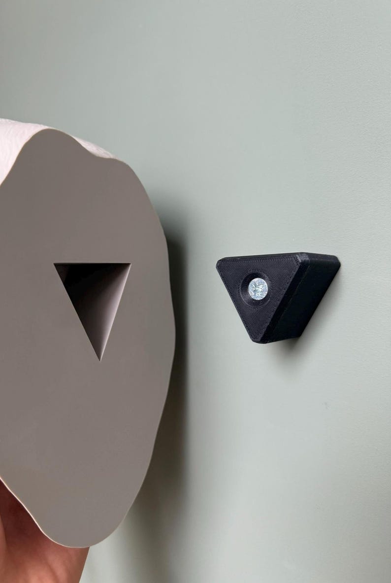 May include: A close-up of a gray, abstract-shaped object with a triangular cutout, next to a black, triangular object with a screw in the center. Both objects are mounted on a light green wall.