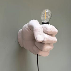 Puede incluir: Aplique de pared con forma de mano de yeso blanco que sostiene una bombilla transparente con una base negra. La lámpara está montada en una pared verde claro, con un cable eléctrico negro que se extiende hacia abajo.
