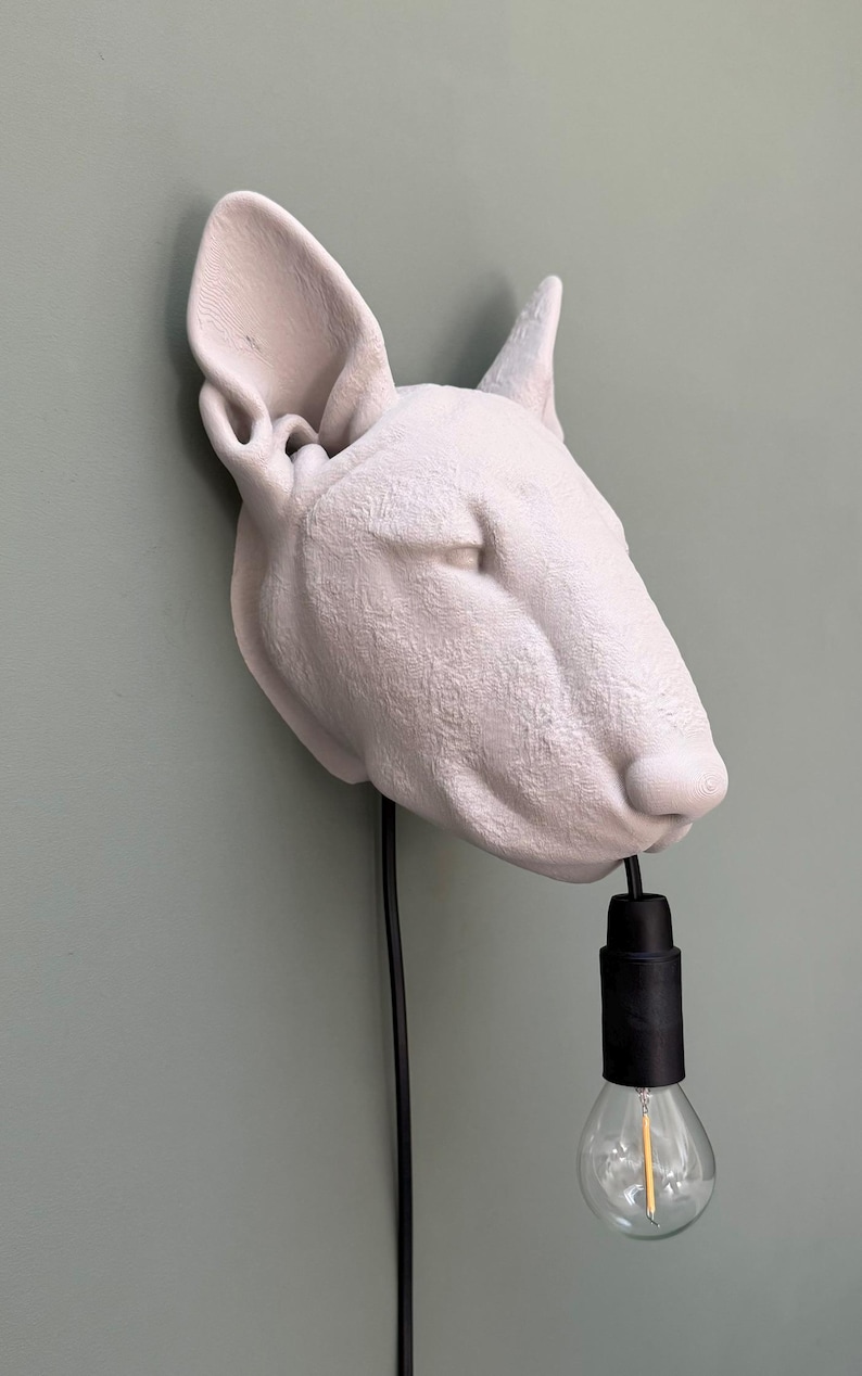 May include: A white Bull Terrier head sculpture with a black cord and lightbulb hanging from its mouth. The sculpture is mounted on a light green wall. The lightbulb is clear with a yellow filament.