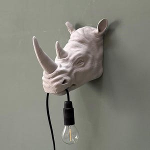 May include: A light gray rhino head wall lamp with a black cord and a clear light bulb. The rhino head is mounted on a light green wall. The lamp is a unique decorative item.