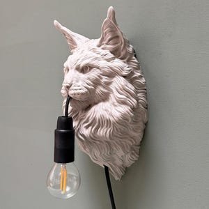 Può includere: Una lampada da parete bianca scolpita a forma di testa di lince. La lampada presenta un portalampada nero e una lampadina trasparente con filamento visibile. La testa di lince è montata su una parete verde chiaro.
