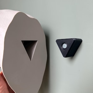 May include: A light gray, organic-shaped toilet paper holder with a triangular cutout, paired with a small black triangular wall mount. The holder is mounted on a light green wall.