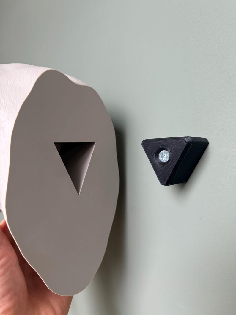 May include: A light gray, organic-shaped toilet paper holder with a triangular cutout, paired with a small black triangular wall mount. The holder is mounted on a light green wall.