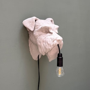 May include: A wall-mounted light fixture shaped like a Schnauzer dog head in a matte off-white finish. The sculpture features detailed fur texture and holds a black electrical cord with a black socket and a clear glass Edison-style light bulb.