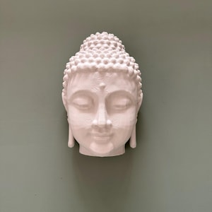 May include: A white Buddha head sculpture with closed eyes and a serene expression. The head features detailed textures on the hair and face, and is mounted on a muted green background. The sculpture is a representation of peace and meditation.