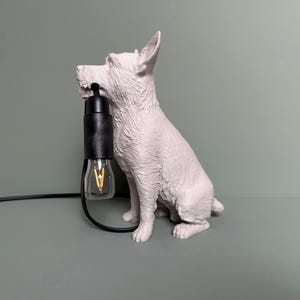 Puede incluir: Lámpara con forma de perro gris claro con un portalámparas negro y una bombilla transparente. El perro está sentado erguido con la boca sujetando el portalámparas. La lámpara tiene un cable negro.