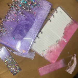 Puede incluir: Un cuaderno morado y rosa con purpurina, cubierta transparente y encuadernación en espiral. El cuaderno tiene un borla y el texto "Do what you love" con un corazón. Viene con dos marcadores a juego.