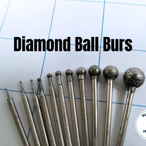 May include: A collection of diamond ball burs of varying sizes, arranged on a grid-lined surface. The burs have silver-colored shafts and diamond-coated ball tips. The text "Diamond Ball Burs" is displayed above the tools.