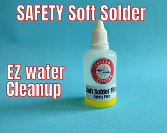 Op de afbeelding: Een doorzichtige fles met zachte soldeerflux met een geel etiket waarop "Soft Solder Flux Safety Flux" staat. De fles is gemarkeerd met een rode cirkel waarop "Lustro Electro" staat en bevat een bliksemflits en een vuist in de cirkel. De fles staat op een lichtblauwe achtergrond.