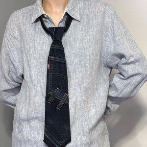 K&ouml;nnte beinhalten: Eine dunkelblaue Jeans-Krawatte mit einer Tasche und dem Markenetikett von Levi's. Die Krawatte hat ein einzigartiges Design, bei dem das Jeansmaterial geschnitten und gen&auml;ht wurde, um ein stilvolles Accessoire zu schaffen. Die Krawatte wird &uuml;ber einem hellblauen Button-Down-Hemd getragen.