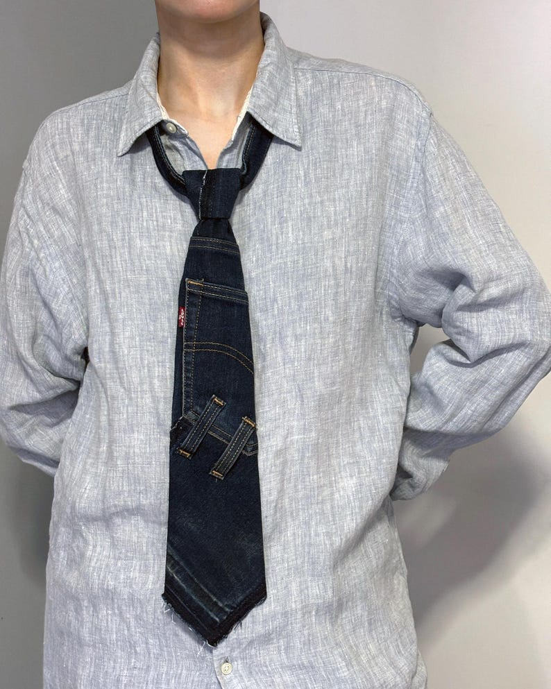 K&ouml;nnte beinhalten: Eine dunkelblaue Jeans-Krawatte mit einer Tasche und dem Markenetikett von Levi's. Die Krawatte hat ein einzigartiges Design, bei dem das Jeansmaterial geschnitten und gen&auml;ht wurde, um ein stilvolles Accessoire zu schaffen. Die Krawatte wird &uuml;ber einem hellblauen Button-Down-Hemd getragen.
