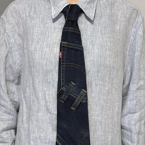 K&ouml;nnte beinhalten: Dunkelblaue Jeans-Krawatte mit Used-Look. Die Krawatte hat eine kleine Tasche und dekorative N&auml;hte, mit zwei Jeansschlaufen unten. Ein rotes Levi's-Etikett ist sichtbar.