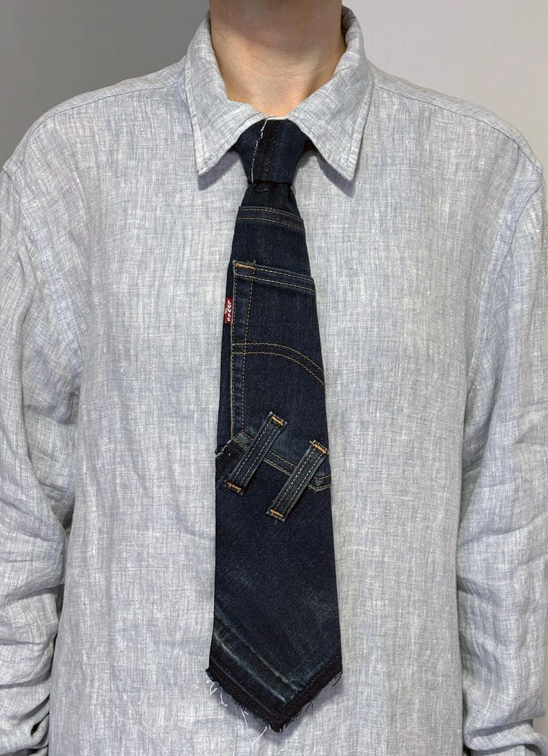 K&ouml;nnte beinhalten: Dunkelblaue Jeans-Krawatte mit Used-Look. Die Krawatte hat eine kleine Tasche und dekorative N&auml;hte, mit zwei Jeansschlaufen unten. Ein rotes Levi's-Etikett ist sichtbar.
