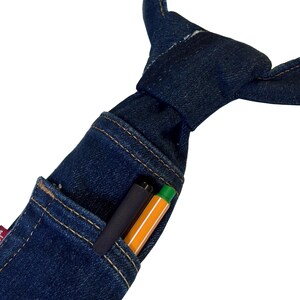 K&ouml;nnte beinhalten: Eine dunkelblaue Jeans-Krawatte mit einer Tasche, die einen schwarzen Stift und einen orangefarbenen Textmarker enth&auml;lt. Die Krawatte ist aus Jeansstoff gefertigt und hat einen klassischen Knoten. Die Tasche ist mit einem Kontrastfaden gen&auml;ht.