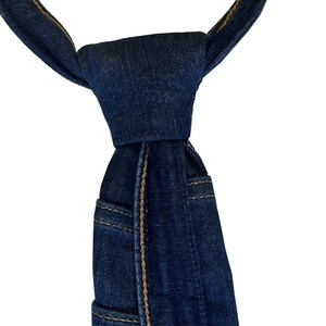 K&ouml;nnte beinhalten: Eine dunkelblaue Jeans-Krawatte mit sichtbaren N&auml;hten und Taschen-Details. Die Krawatte ist oben geknotet und zeigt die Textur und Konstruktion des Jeansstoffs. Das Design ahmt das Aussehen einer Jeans nach.